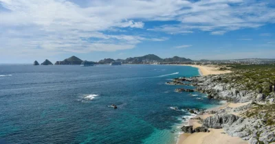 Aerial view of the Los Cabos coastline with turquoise waters, sandy beaches, and the city skyline stretching along the shore in Baja California Sur.